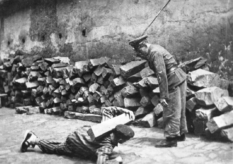 An SS soldier in a camp, watching an inmate crawling in front of him carying a block of stone on his shoulders.